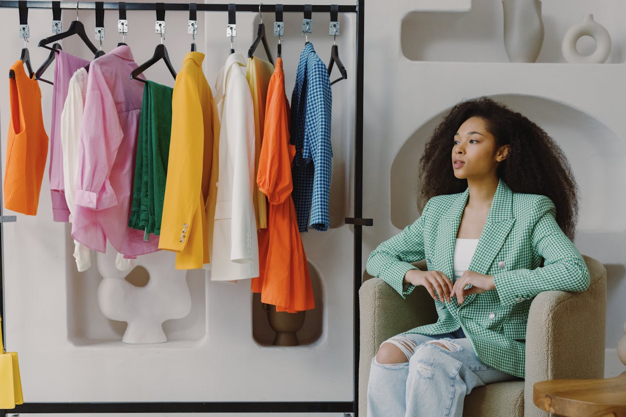 Stylish woman in green blazer sitting by colorful clothes in boutique.
